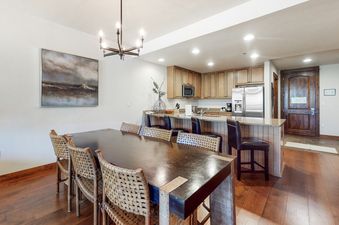 The dining area in BlueSky Breckenridge 504 features a long wooden table with six woven chairs, illuminated by an overhead chandelier, and a spacious adjacent kitchen with a breakfast bar.