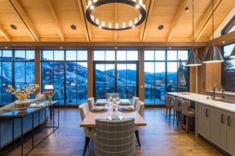 A modern dining room with a large wooden table and chairs, overlooking a snowy mountain range from floor-to-ceiling windows.