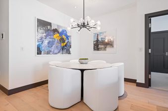 A modern dining area featuring a round marble table, four white chairs, and a contemporary chandelier, set against white walls adorned with two art pieces.