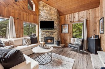 A cozy living room in The Village House with an L-shaped sofa, stone fireplace, mounted TV, and large windows looking out onto a green forest.