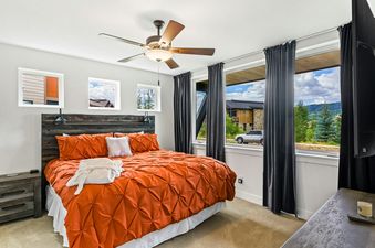 A bedroom with a king-size bed with an orange comforter, a wooden headboard, and two windows with a view of a mountain landscape.