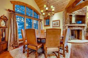 A rustic dining room with a large wooden table and leather chairs next to a window overlooking a snowy forest.