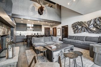 A modern rustic open-concept living area and kitchen at Snow Cloud Lodge 506 with grey sofas, a stone fireplace, and a prominent moose head decoration.