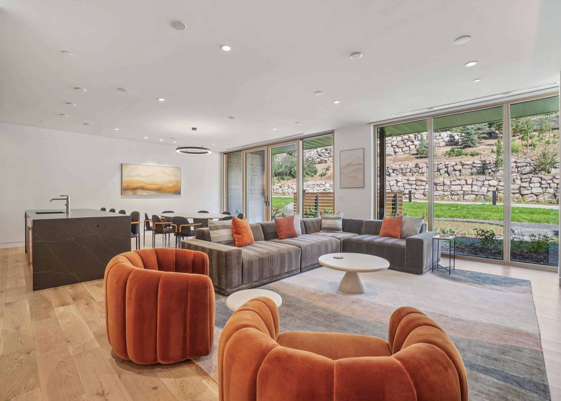 A modern living room with a large grey sectional, orange armchairs, a white coffee table, and expansive windows offering a view of outdoor rock walls and greenery.