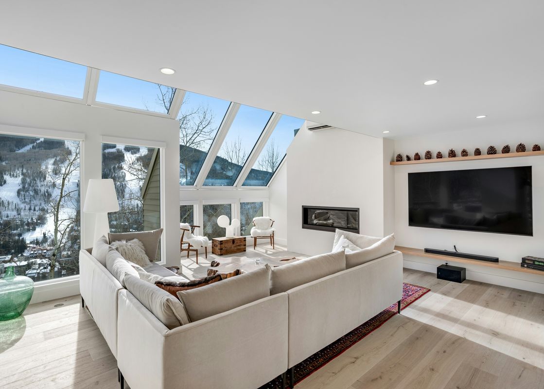A modern and bright living room at Luxe Lookout featuring a large sectional sofa, a sleek fireplace, a flat-screen TV, and expansive windows with views of snow-covered mountains.