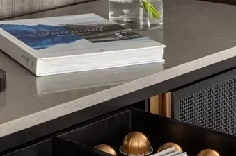A drawer filled with coffee capsules, napkins, and spoons, with a book and flowers on the counter above, showcasing the amenities of the Fairmont Banff Springs Gatehouse 2 Queens.