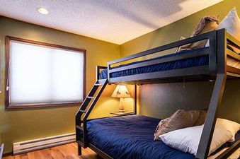 A cozy bedroom in Ranch at Steamboat RA106 featuring a black metal bunk bed with blue comforters, a warm table lamp, and a large window with a white blind.