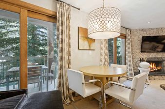 A modern, well-furnished living room with a fireplace, a round dining table, and a window with a view of the forest outside.