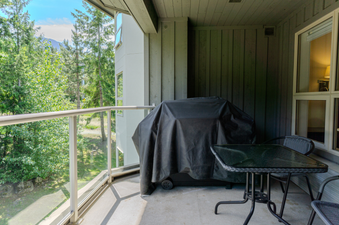 A spacious balcony at Aspens on Blackcomb Unit 451, featuring a grill covered in black, a glass-top table with two chairs, and a view of lush green trees and mountains under a blue sky.