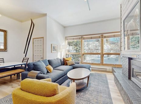 Bright living room in Kutuk 108 with a blue sectional sofa, yellow armchair, fireplace, and large windows showing a snowy landscape.