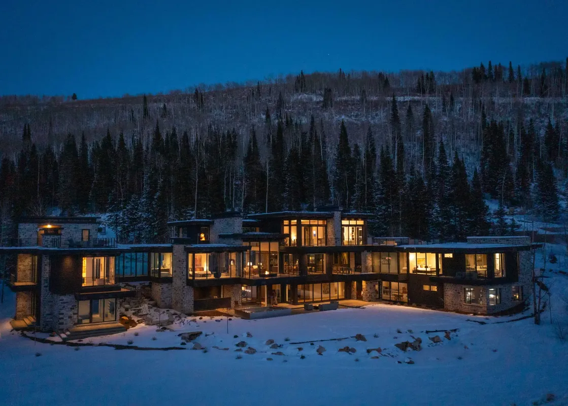 White Pine Canyon is a luxurious, modern home nestled in the snowy woods. The exterior features large windows that provide stunning views of the surrounding nature, with a snowy yard and trees in the background.