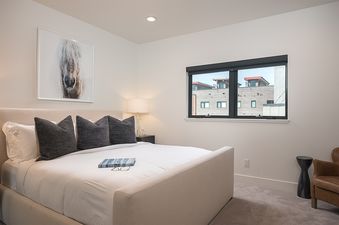 A comfortable bedroom at Princetown Grande features a cream upholstered bed, a framed horse art piece, and a window with a view of other modern buildings.