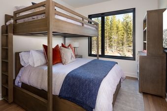 A cozy bedroom with a bunk bed, a large window with a view of a pine forest, and a wooden cabinet.