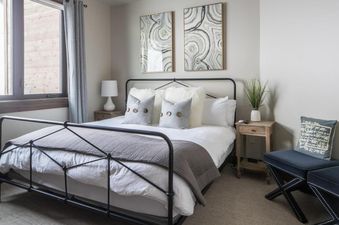 A cozy bedroom with a queen-sized bed, white linens, gray throw, and abstract art, featuring a window and nightstands.