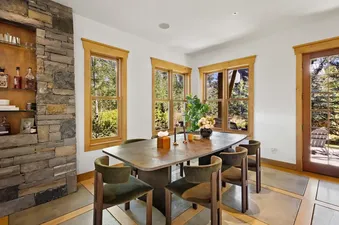 A rustic dining room with a long wooden table, green upholstered chairs, a stone accent wall with shelves, and multiple windows looking out to a lush green forest.