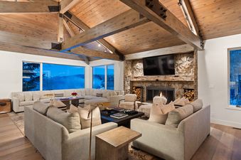 A luxurious living room with vaulted wooden ceilings, a large stone fireplace, and expansive windows offering stunning views of a snowy mountain landscape.