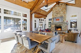 The magnificent great room at Snow Dance at The Colony with a rustic dining table, large stone fireplace, and cathedral wooden ceilings overlooking a snowy landscape.