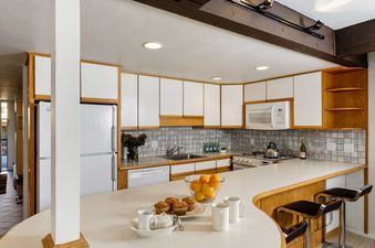 A bright and functional kitchen in Aspen Alps Apartment 101 features white cabinetry with natural wood trim, a curved breakfast bar with stools, and modern appliances.