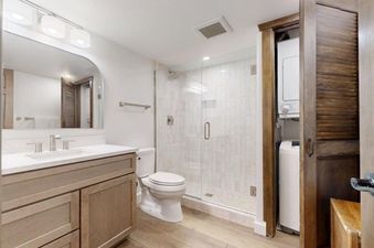 A modern bathroom at Slope 101 with a vanity, toilet, glass-enclosed shower, and a stacked washer/dryer hidden behind a louvered door.