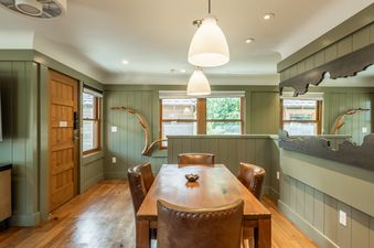 A cozy dining area featuring a wooden table, leather chairs, green paneled walls, and large windows with outdoor views.