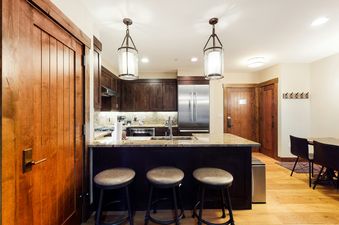 A modern kitchen with dark wood cabinets, light granite countertops, stainless steel appliances, a kitchen island with bar stools, and warm lighting.