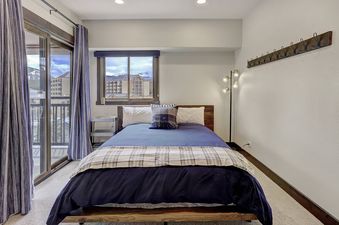 A bedroom with a king-sized bed, a dresser, a lamp, a window with a view of the mountains, and a door to a balcony.