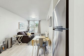 A bright and airy living room at Big Sky Mountain View Escape, featuring a brown sofa, a dining table with chairs, and a stainless steel refrigerator, all under natural light from a large window.