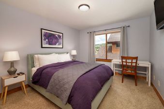 A serene bedroom at 2002 Lupine Lane features a comfortable bed with a purple duvet, a white desk, and a window offering a mountain view.