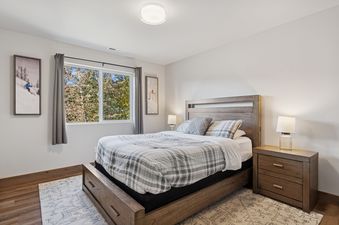 A cozy guest bedroom with a plaid duvet, wooden furniture, and a window overlooking trees.