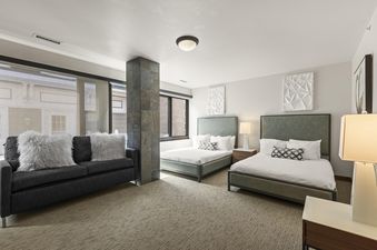 A modern bedroom in The Lowell 401 with two twin beds featuring green headboards, a dark gray sofa with fuzzy white pillows, and large windows.