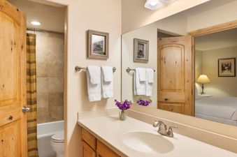 A modern bathroom at Montaneros 314 features a vanity with a single sink, a large mirror, and a shower-tub combination, with a glimpse of the adjacent bedroom.