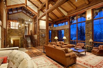 A spacious and luxurious living area at Treehouse on West Fork, featuring high ceilings with exposed timber beams, comfortable brown sofas, leather armchairs, a coffee table, and large windows offering views of a snowy outdoor landscape.