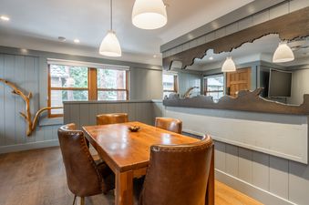 A cozy dining area at Cedar Crest Stellars Jay Cottage 4 features a wooden table, brown leather chairs, and custom-cut wooden mirror, illuminated by pendant lights and offering views of a snowy landscape.