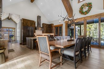A spacious dining room and kitchen with a large wooden table, modern chandeliers, and a view of trees outside.