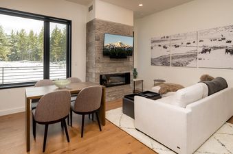A cozy living room in 230 LakeLodge with a dining area, a modern fireplace, a flat-screen TV, and a white sofa, featuring large windows with winter forest views.