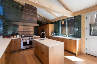 A modern kitchen in Galloping Goose Chalet featuring light wood cabinets, dark green tiled walls, and a central island with a sink.