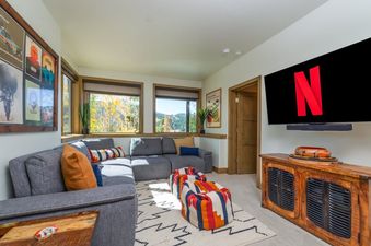 A cozy entertainment area with a large gray sectional sofa, colorful poufs, and a wide-screen TV displaying the Netflix logo.