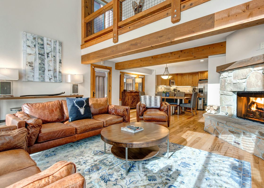 The open-concept living area at Deer Valley 355 B features brown leather seating, a stone fireplace, and exposed wooden beams, seamlessly connecting to the dining and kitchen areas.