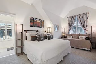 A bedroom with a king-sized bed with white bedding, a white bed skirt, and several pillows. There is a side table with a lamp next to the bed, and a large window with curtains in the background.