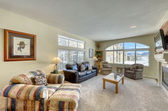 A living room with a large window overlooking a snowy mountain range, with a fireplace to the right, a leather couch, a couch with a striped ottoman, a coffee table, an armchair and a bookcase with a lamp.