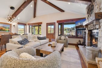 A cozy and elegant living room in the Park Avenue Home, featuring a stone fireplace, plush sofas, a modern chandelier, and large windows with outdoor views.