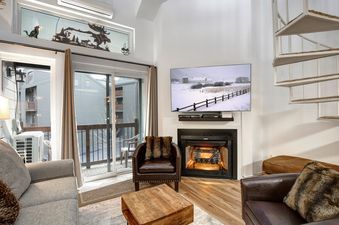 A cozy living room featuring a flat-screen TV above a fireplace, a grey sofa, two brown leather armchairs, a wooden coffee table, and a balcony with a view of a winter landscape.