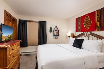 A cozy bedroom at Montaneros 211 featuring a queen-sized bed with white linens, a TV on a wooden dresser, and a vibrant tapestry above the bed.