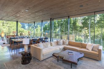 A large living room with a sectional sofa and a coffee table, all facing a large window that looks out over a forest of trees.