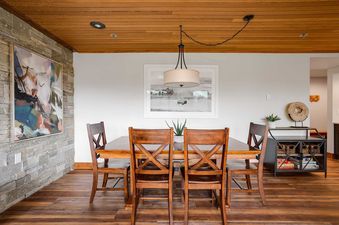 A modern dining area at Chateau Chamonix CX211 with a wooden table and chairs, illuminated by a drum-shade chandelier, and accented by abstract art on a stone wall.
