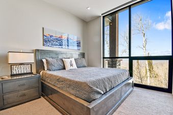 A modern bedroom with a large bed, a nightstand with a lamp, and a full-height window overlooking a mountain landscape.