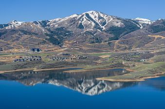 A panoramic view of a snow-capped mountain range, a clear blue lake reflecting the mountains, and a village with residential buildings and a ski resort.