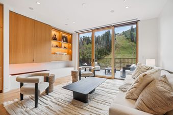 The Founders Place 2401 living area features a cream sofa, modern armchairs, and a sleek coffee table, with a built-in wooden cabinet and large glass doors leading to a balcony with mountain views.