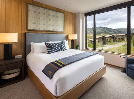 A king-sized bed with white bedding and a striped throw is featured in The Sylvan Lodge King ADA guest room, with a large window offering mountain views.