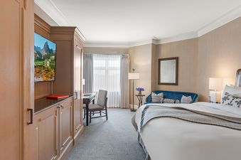 A luxurious bedroom is shown in the St Regis King Room, featuring a large king-sized bed with crisp white linens and a plush gray throw, alongside elegant wooden cabinetry, a flatscreen TV displaying a scenic mountain and lake view, a cozy blue sofa, a sm